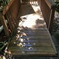 A wooden ramp with railings leading up to a doorway, surrounded by partial shade and foliage.