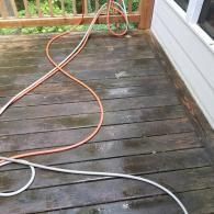 An orange and white garden hose lies across a weathered wooden deck surface next to the side of a house.