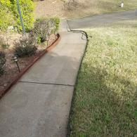 A concrete walkway curves past a landscaped garden bed with reddish edging and a grassy lawn on a sunny day.