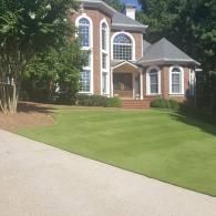 A two-story brick house with a manicured green lawn and concrete driveway under a blue sky.