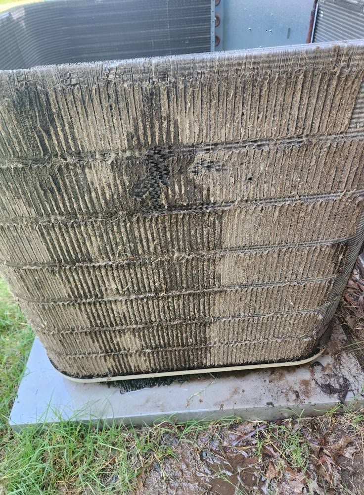 Dirty outdoor HVAC condenser unit with bent, stained fins on a concrete pad beside grass.