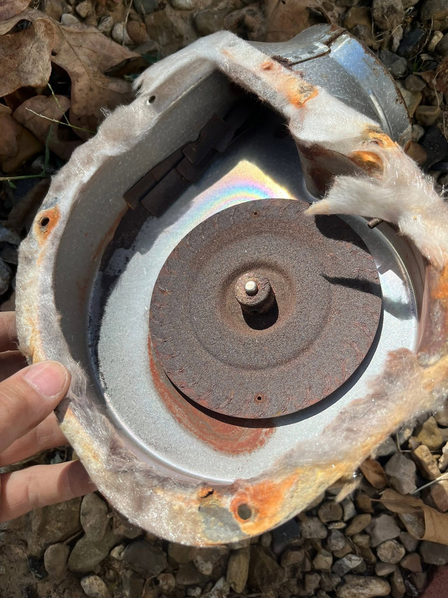 Broken metal speaker cone with rust and exposed circular diaphragm being held outdoors on gravel.