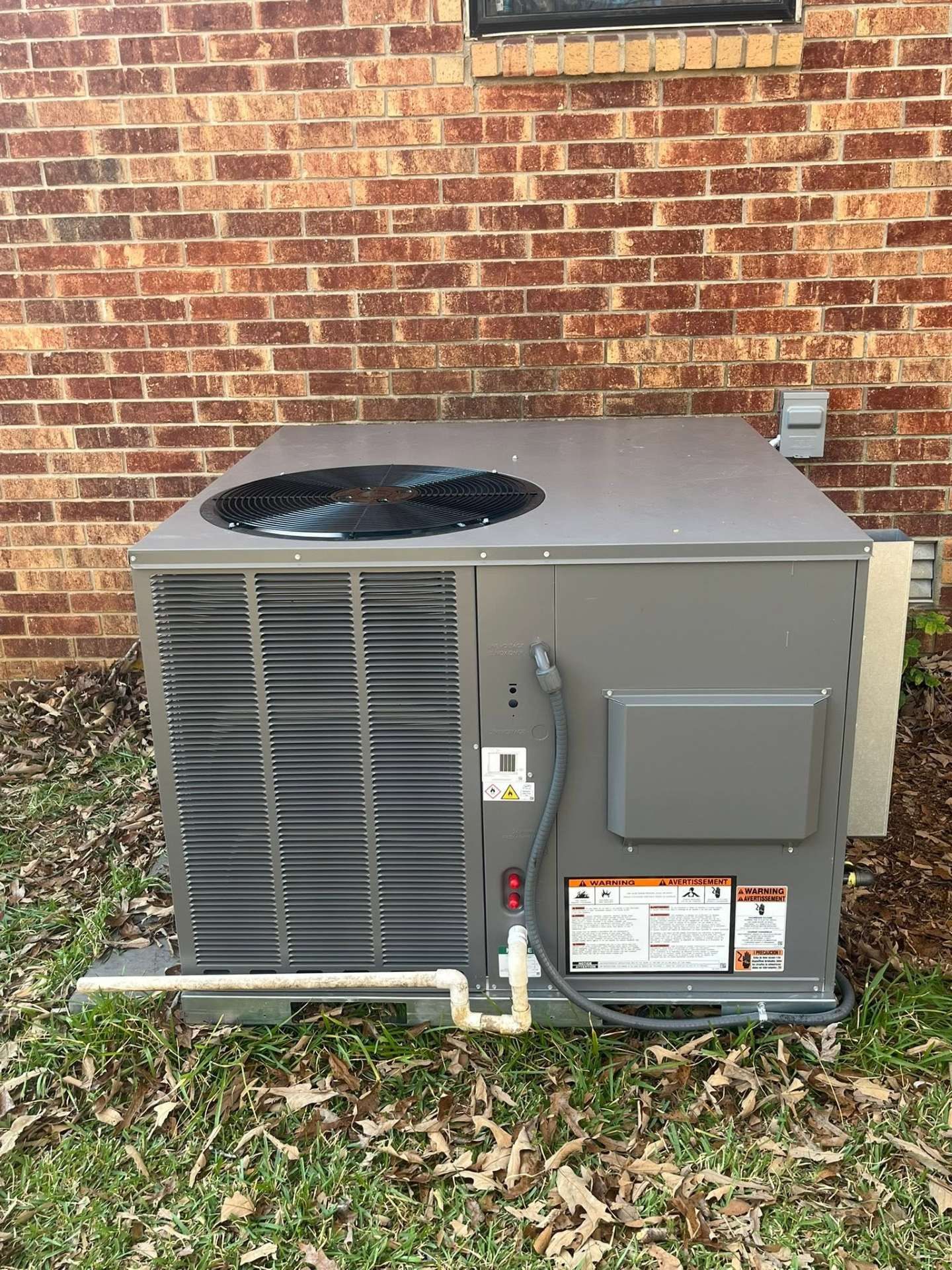Outdoor HVAC condenser unit beside a brick wall, with leaves on the ground and a fan visible on top