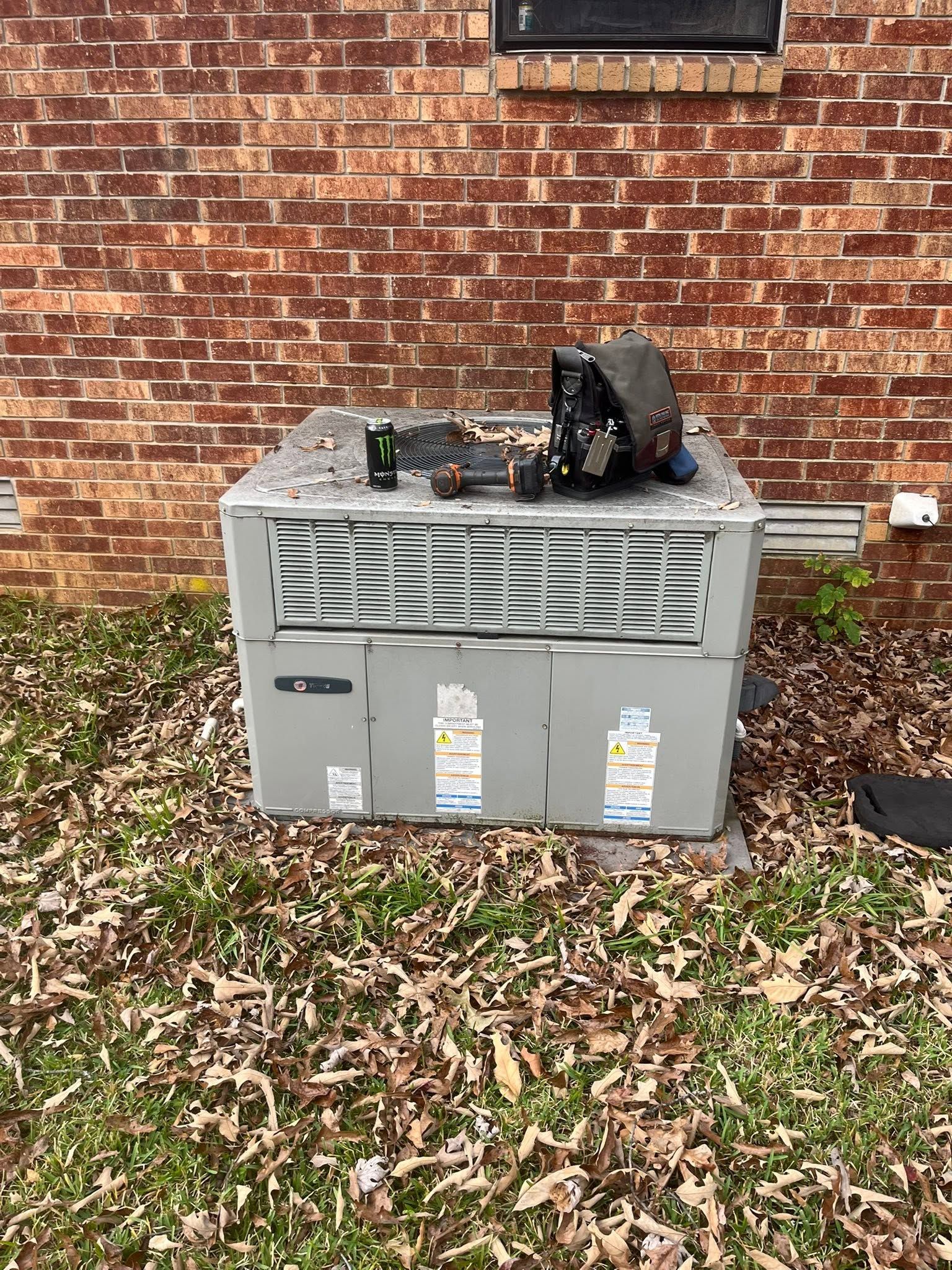 Outdoor HVAC unit with exposed wiring and debris beside a brick wall and fallen leaves