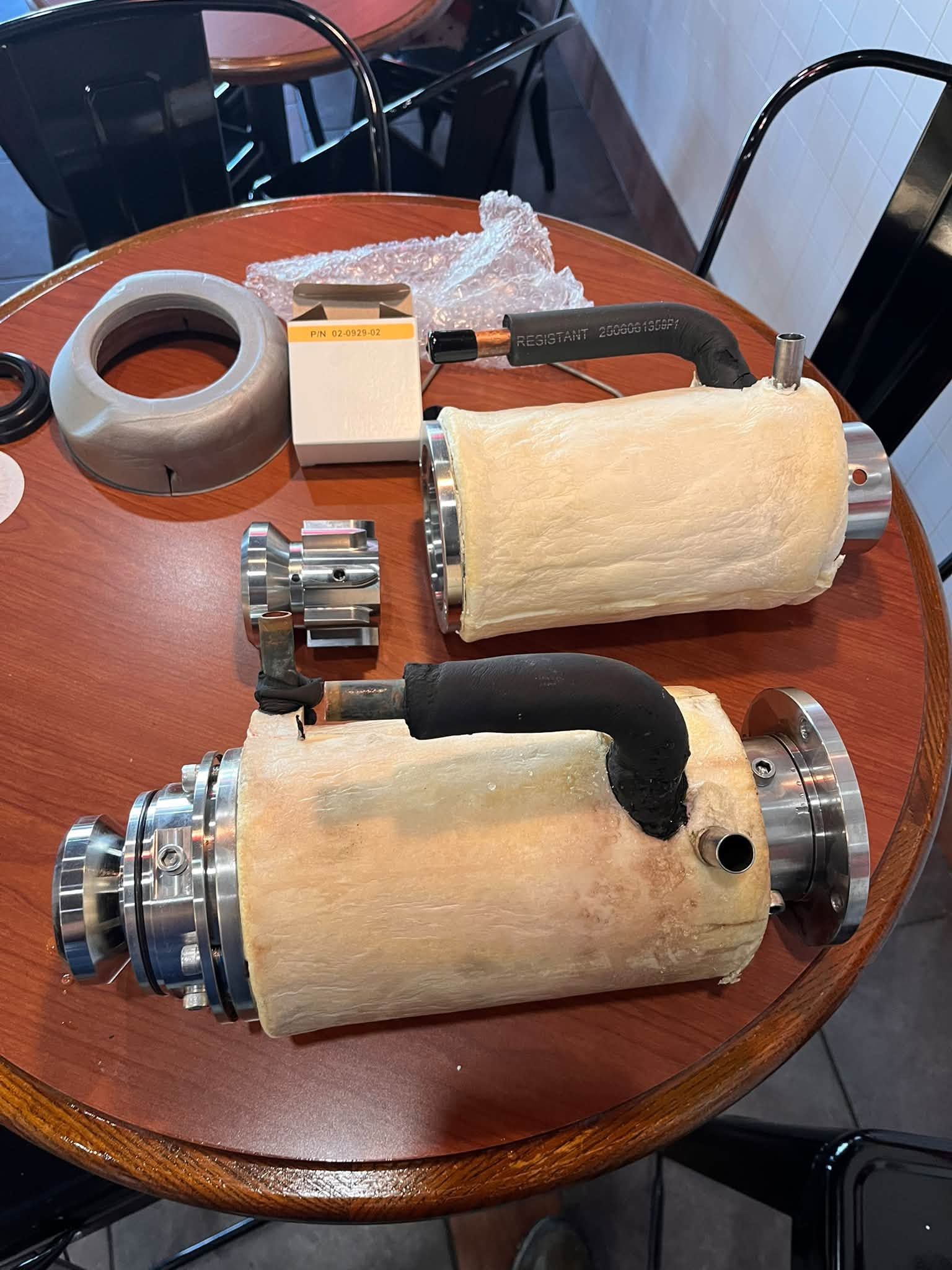 Two white insulated cylindrical parts with metal fittings and hose attached, plus gray tape on a wooden table.