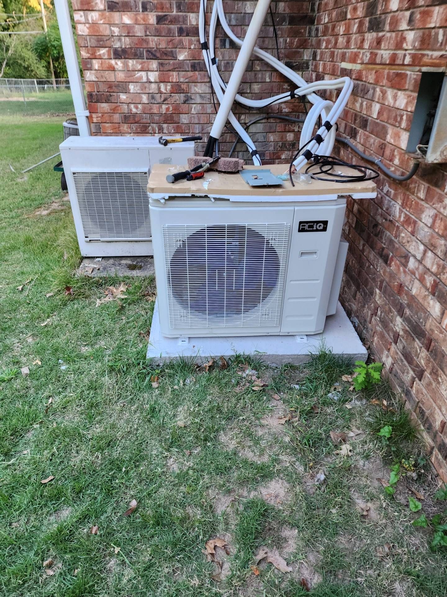 Two outdoor HVAC units beside a brick wall, with exposed pipes and hoses on the ground.