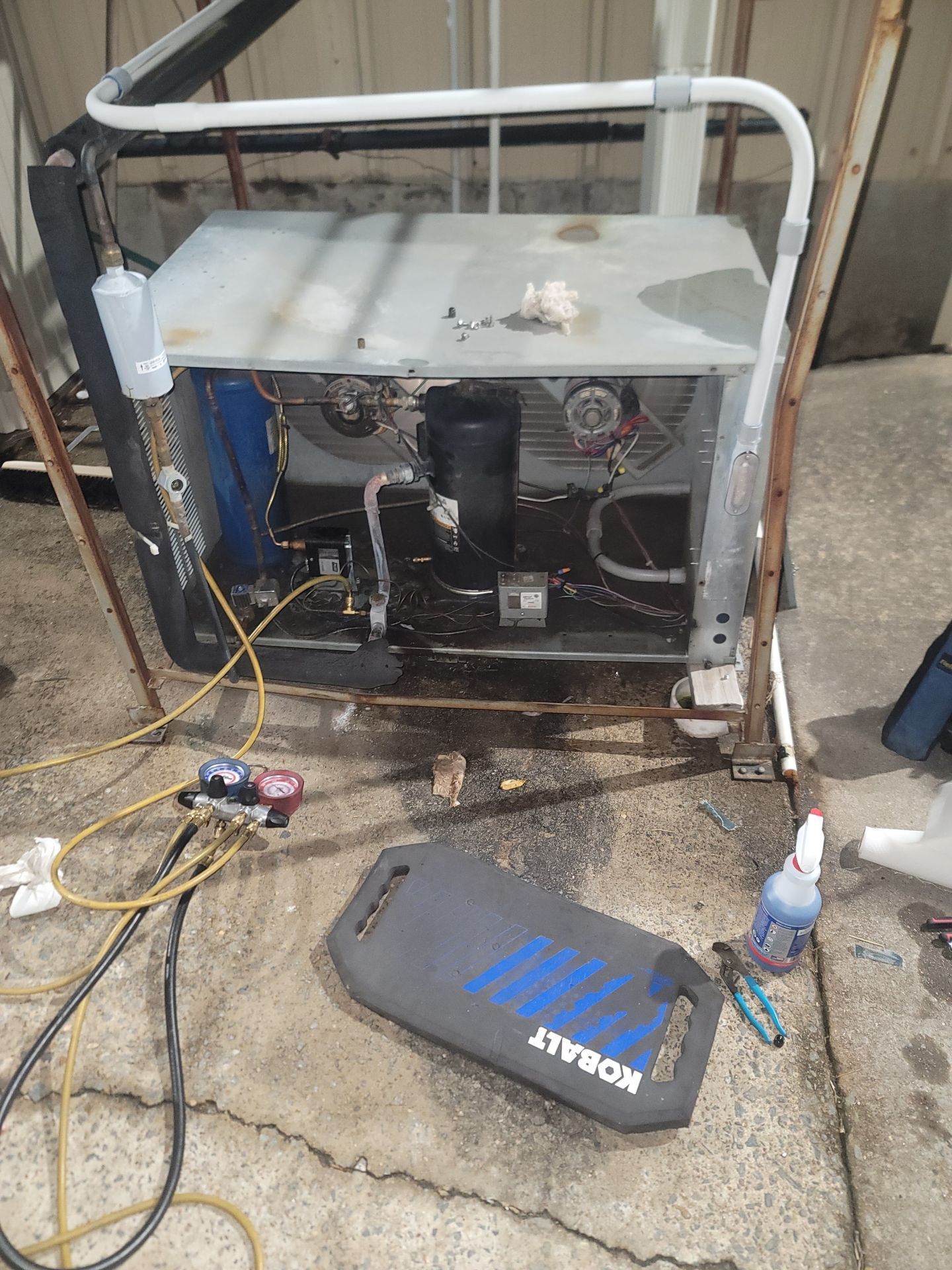 Damaged utility machine with exposed internals on wet concrete, surrounded by hoses and tools.