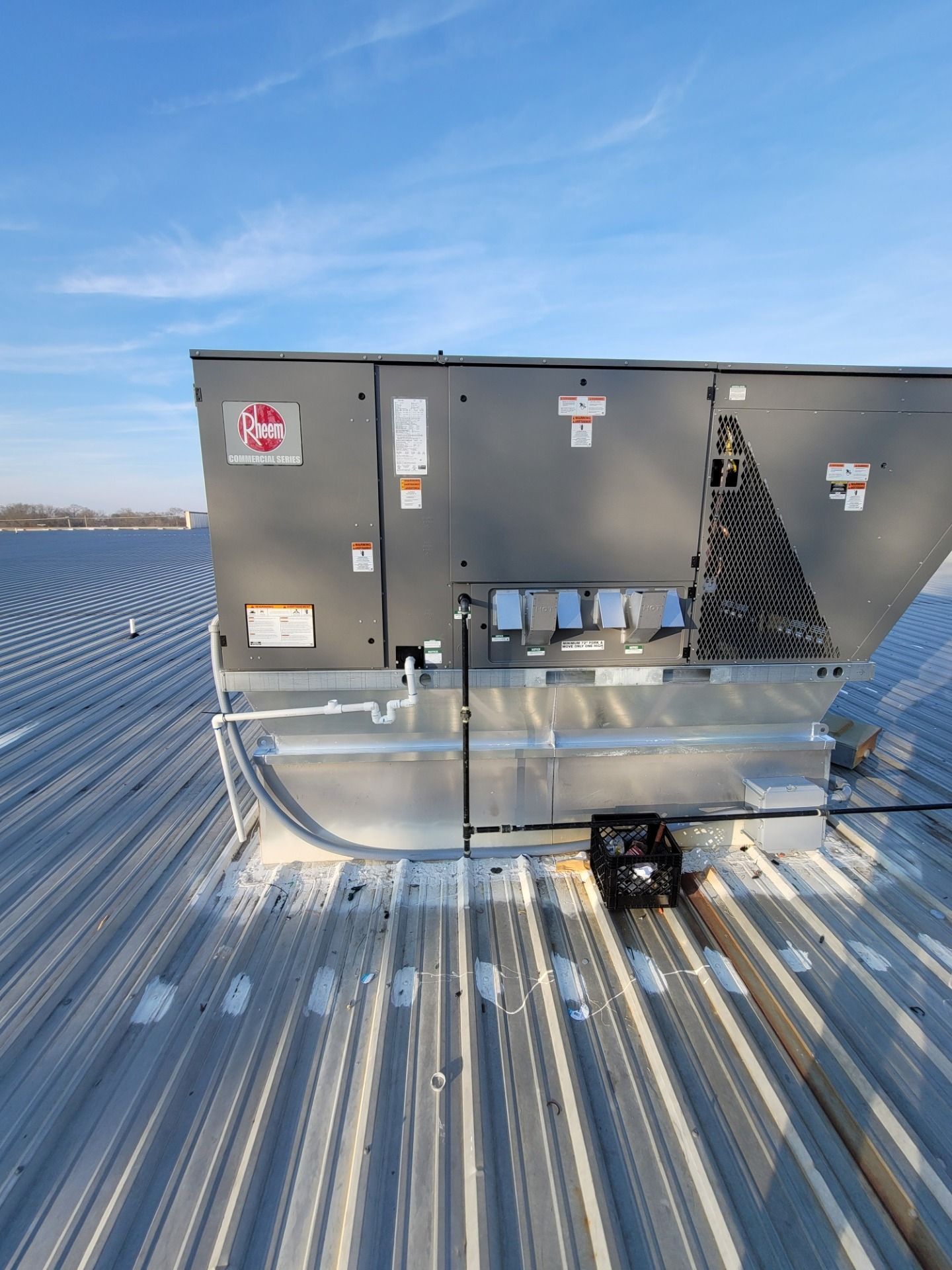 Gray HVAC unit on a metal rooftop, with ducts and a bright winter sky in the background.