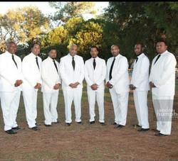 Eight men in matching white suits and black ties stand in a row outdoors on a grassy area, posing for a group portrait.