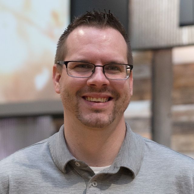 a man wearing glasses and a grey shirt is smiling for the camera .