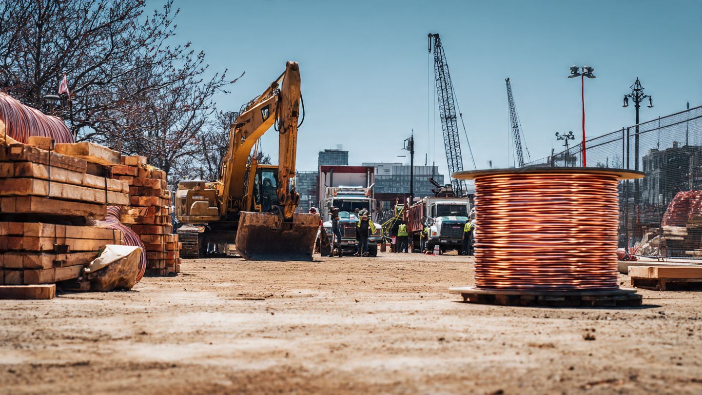 A construction site featuring a yellow excavator, stacks of lumber, and a large spool of copper wire on a dirt ground.