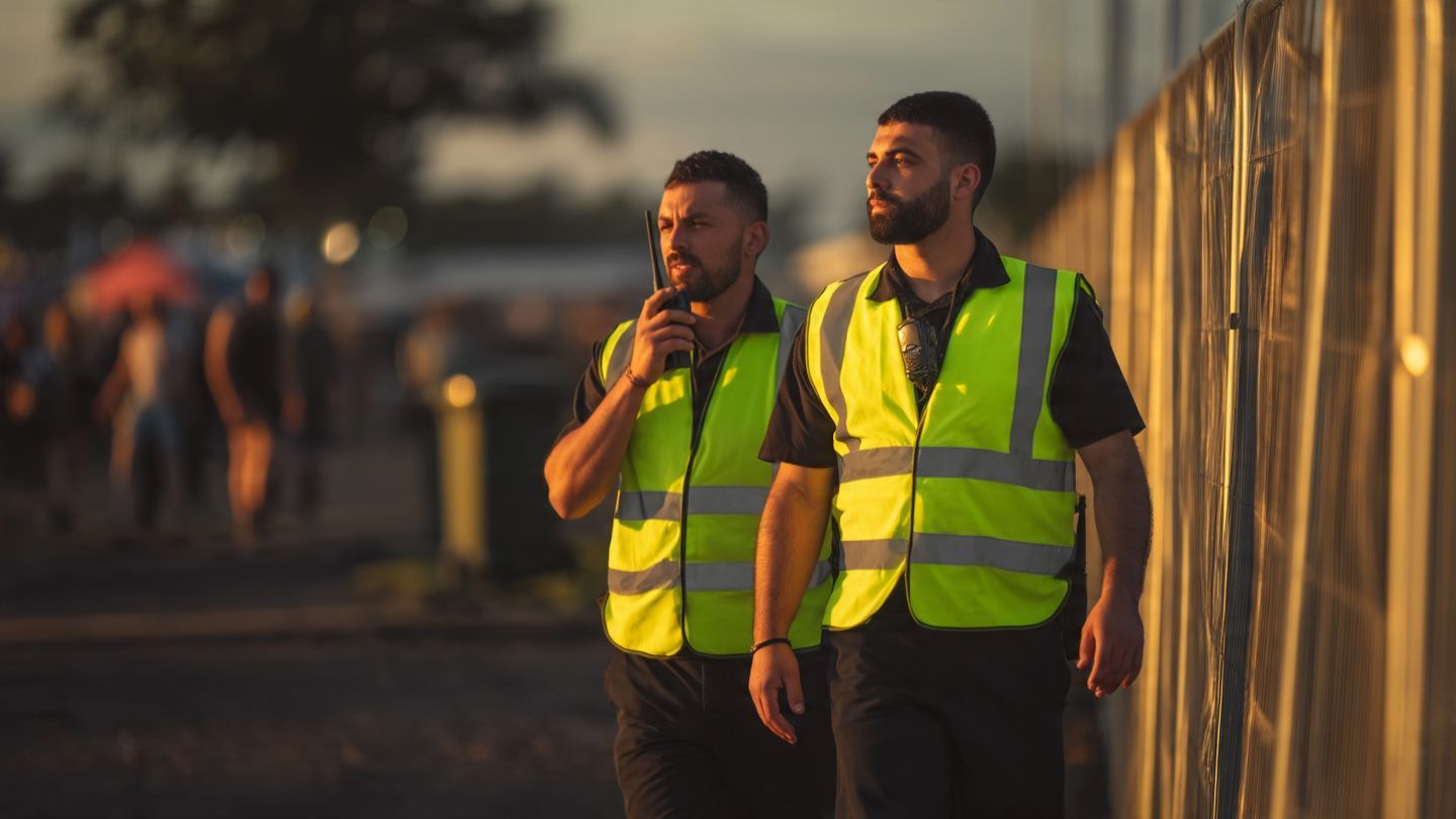 Two workers in neon safety vests standing beside a fence at dusk