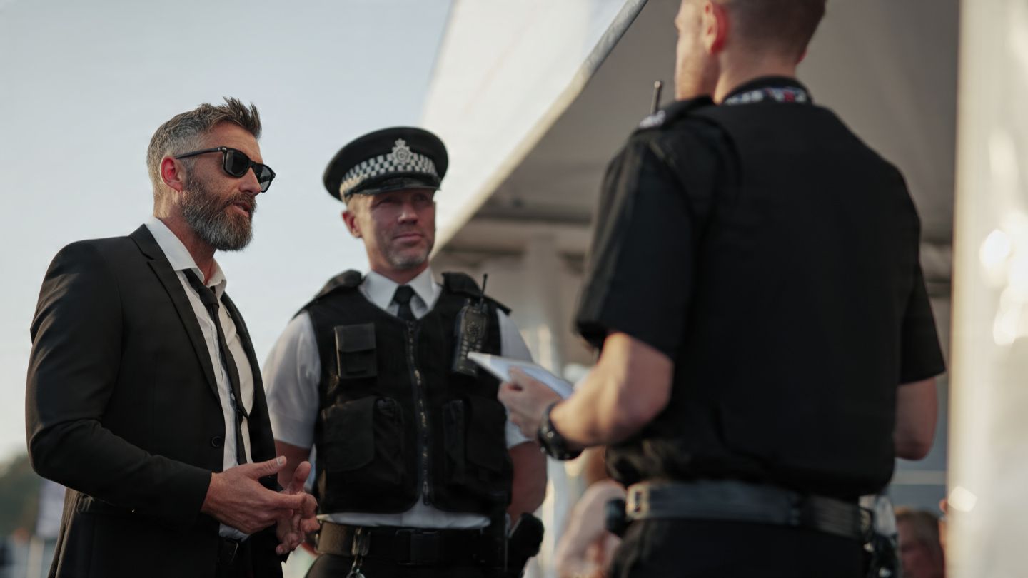 Three men stand talking outdoors, including a police officer in uniform and two men in dark clothing.