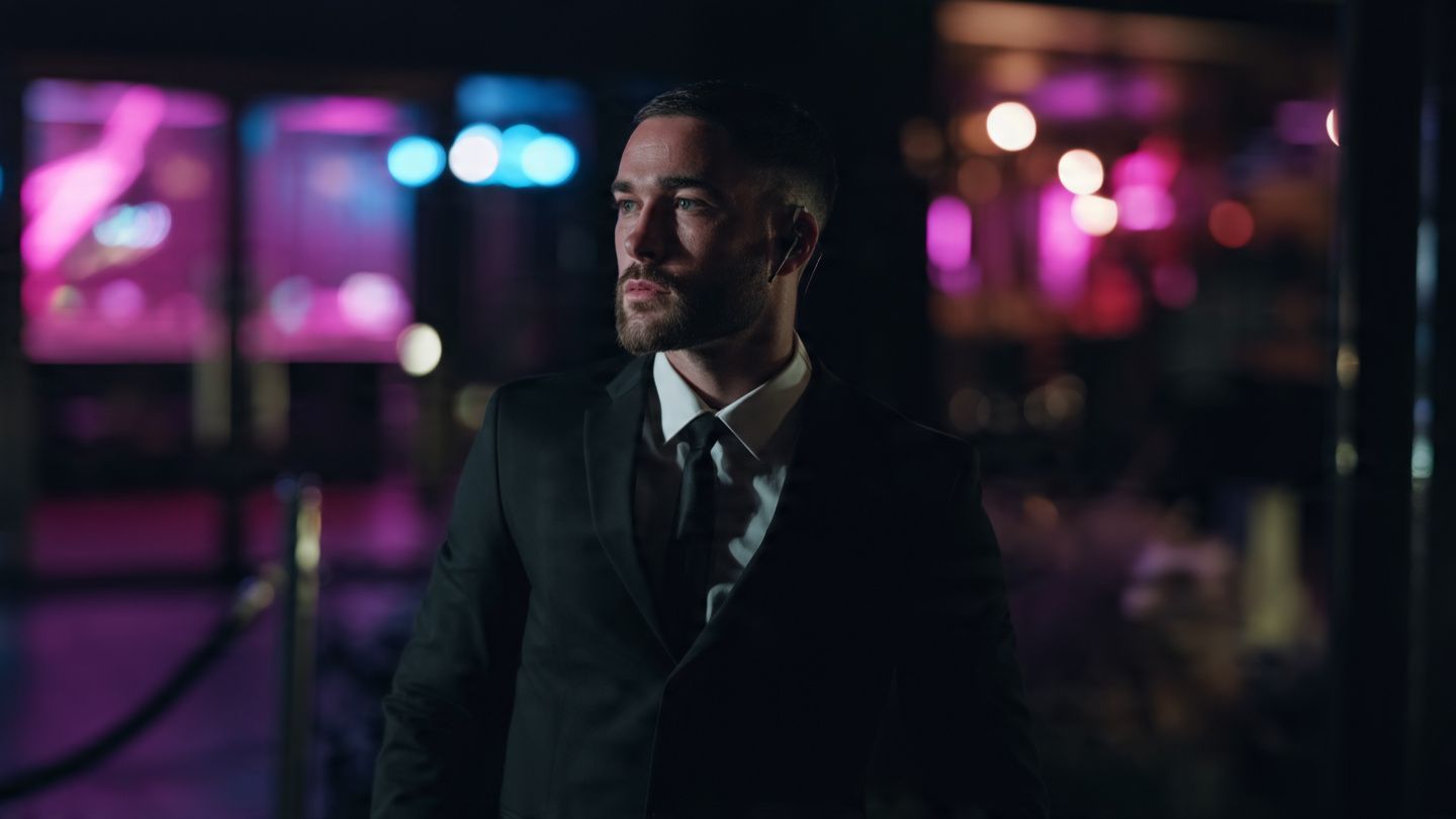 Man in a dark suit standing in a neon-lit bar, looking to the side with a serious expression