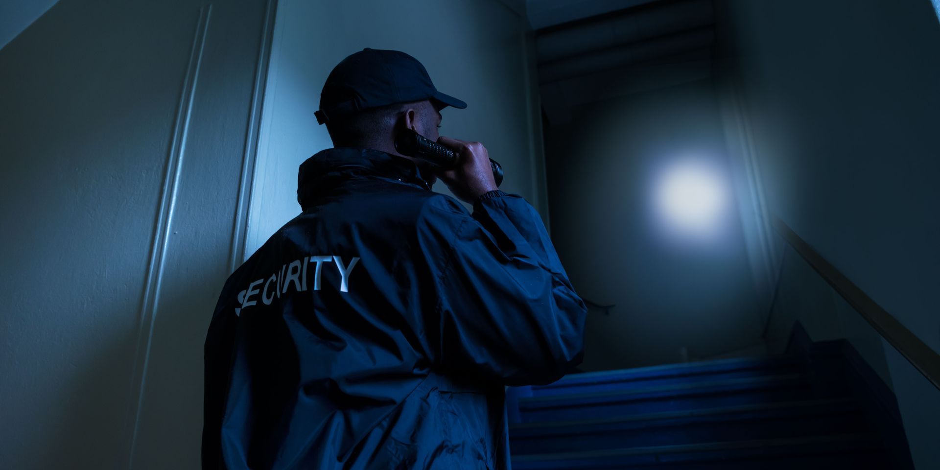 Security guard on a dimly lit staircase, wearing a cap and jacket, talking on a radio.