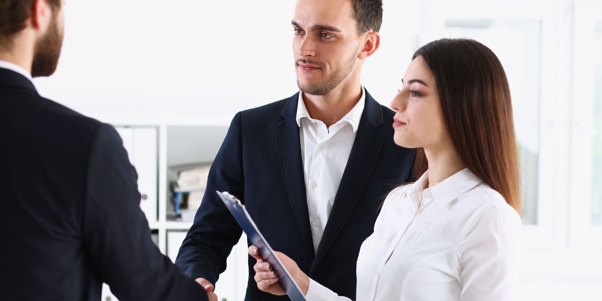 Business people shaking hands in an office setting. A woman looks on holding a clipboard.