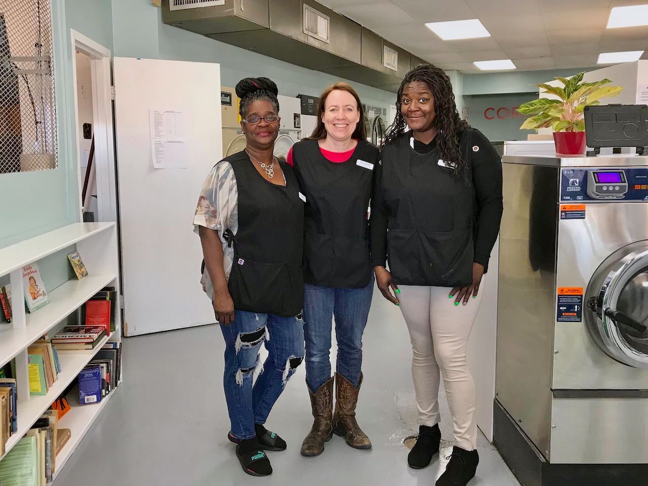 Three women standing together smiling in a laundromat near bookshelf and washing machine