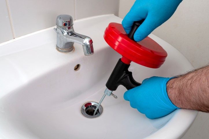 Gloved hands using a red plunger to clear a bathroom sink drain