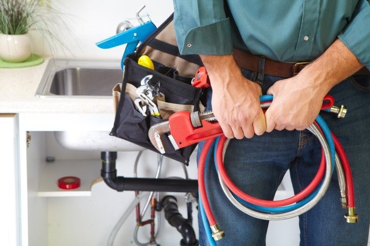 Plumber holding red and blue hoses beside a sink and tool bag under a cabinet
