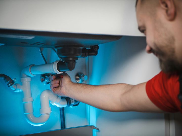 Plumber fixing a sink pipe under a cabinet with a wrench, lit by blue light.