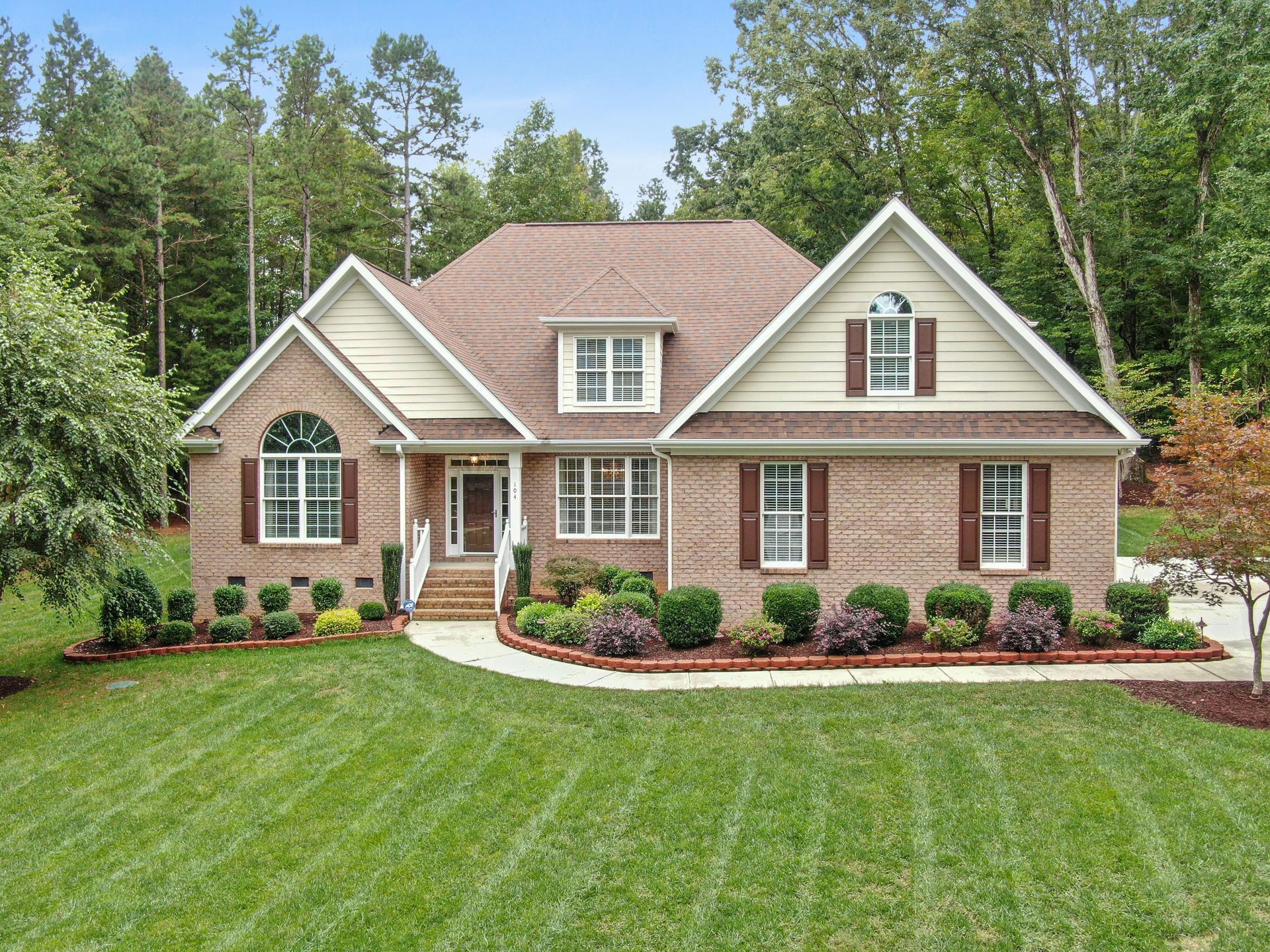 Brick house with brown roof, shutters, and landscaping. Front yard with green grass and trees in the background.