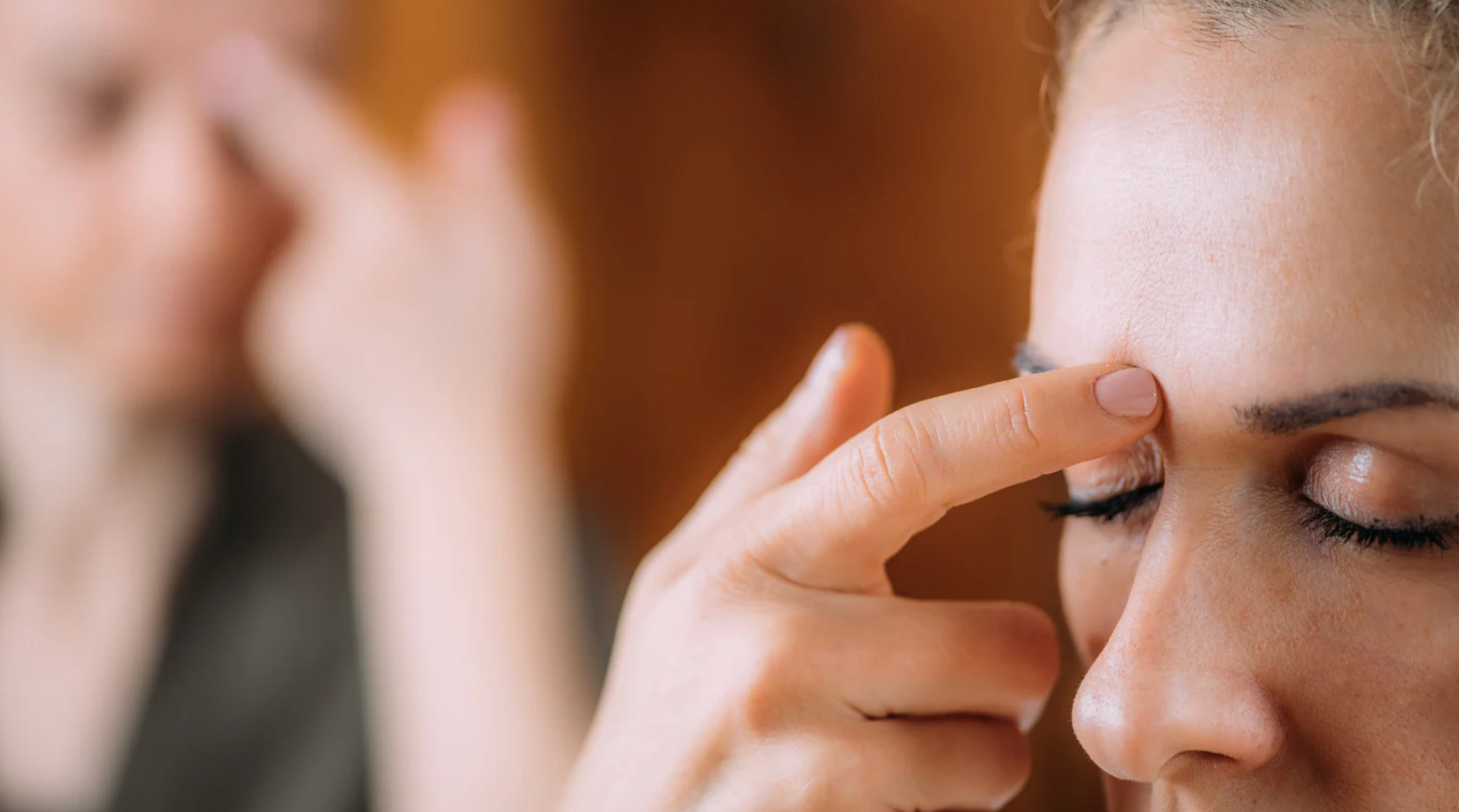 La main d'une personne touche le front d'une autre, entre les sourcils ; les yeux sont fermés. Une personne floue se trouve à l'arrière-plan.