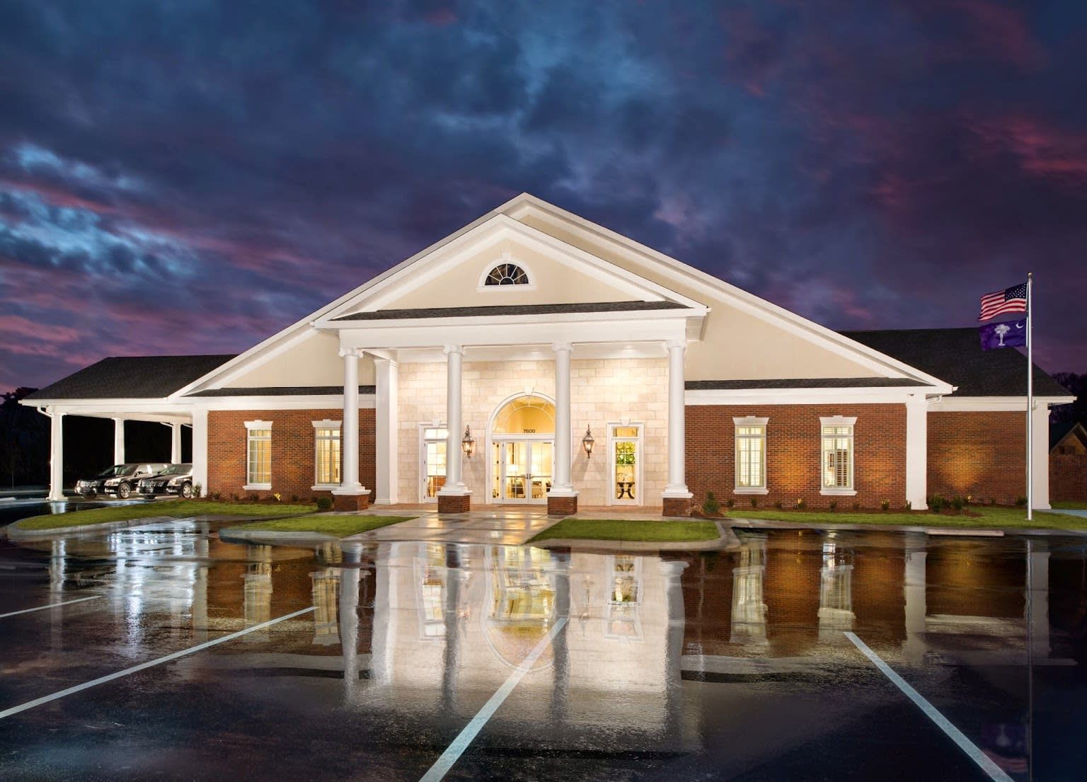 Brick building with white columns and lit entrance reflected on a wet parking lot at dusk