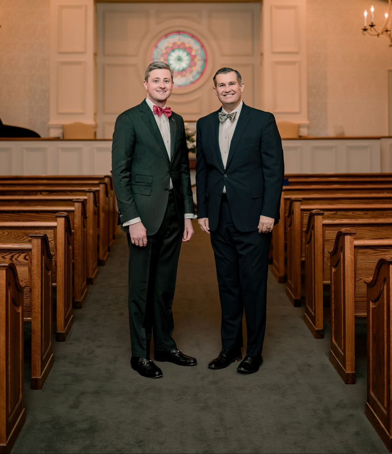 Two men in dark suits standing in a church aisle, smiling under warm light.