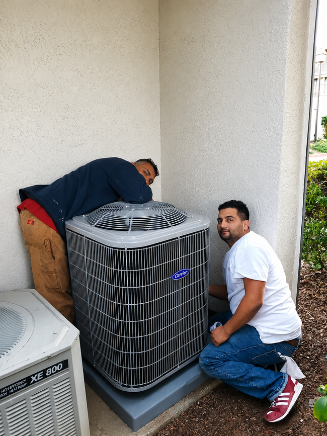 Two men inspecting an outdoor air conditioner unit beside a building wall.