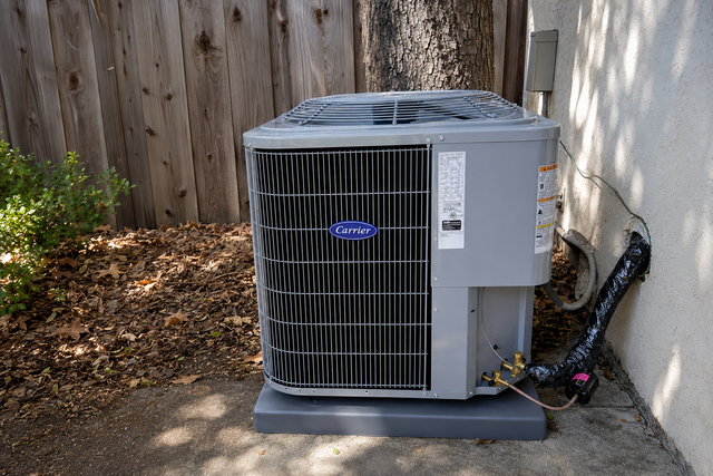 Outdoor HVAC condenser unit beside a house, sitting on a concrete pad near a wooden fence