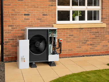 Outdoor heat pump unit beside a brick building wall and window on a paved patio