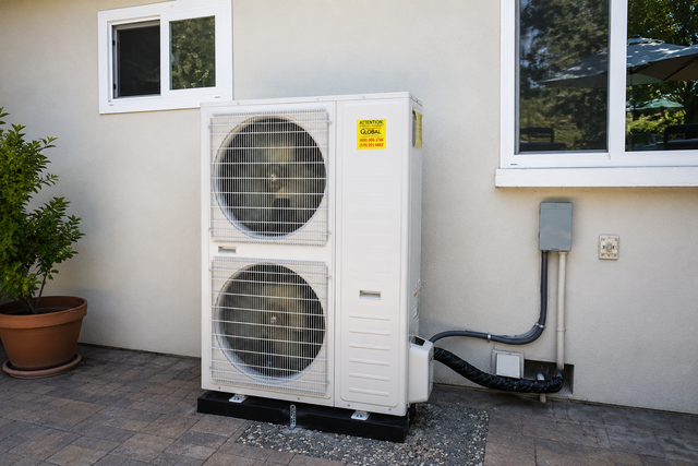 Outdoor heat pump unit mounted beside a house wall with windows and utility lines nearby