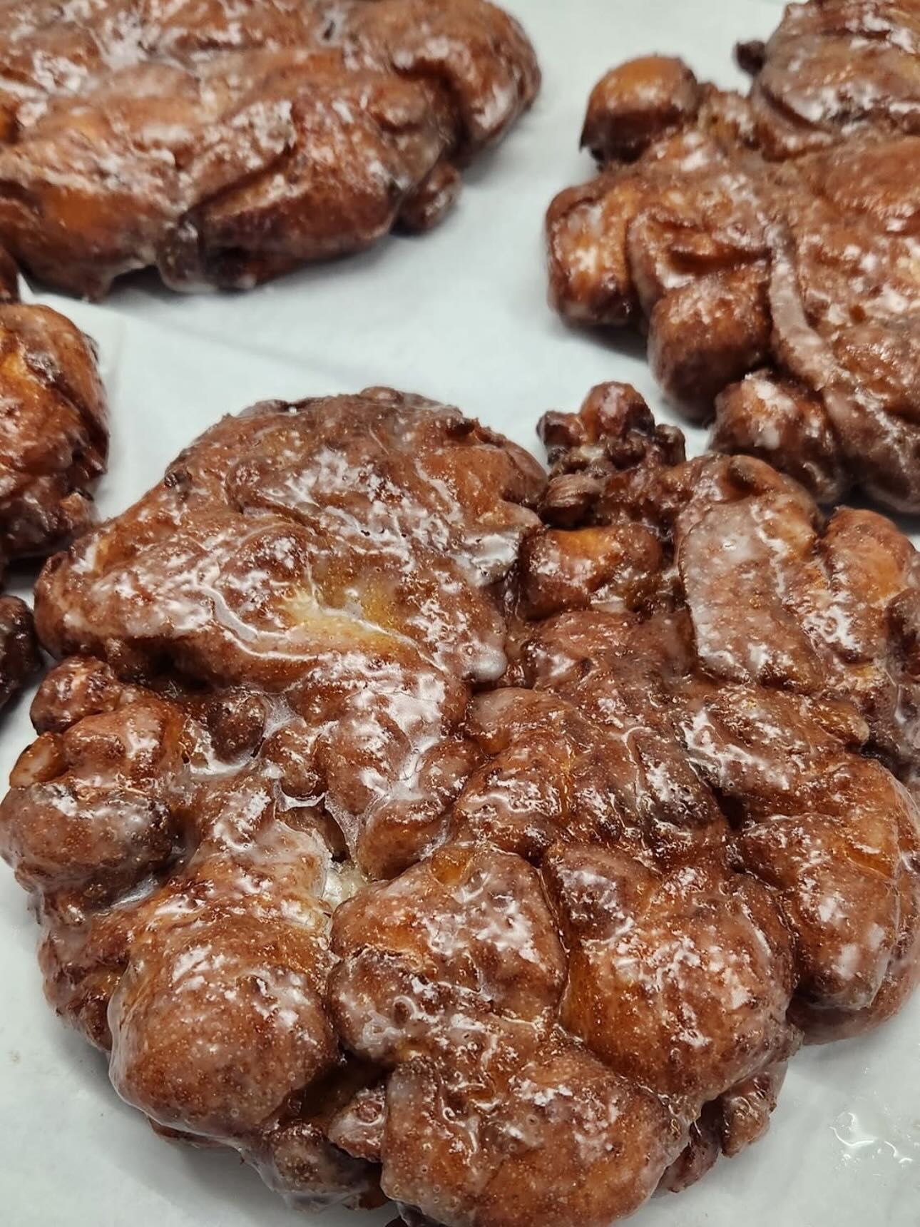 Close-up of glazed apple fritters, various sizes, on parchment paper. Brown, shiny glaze.