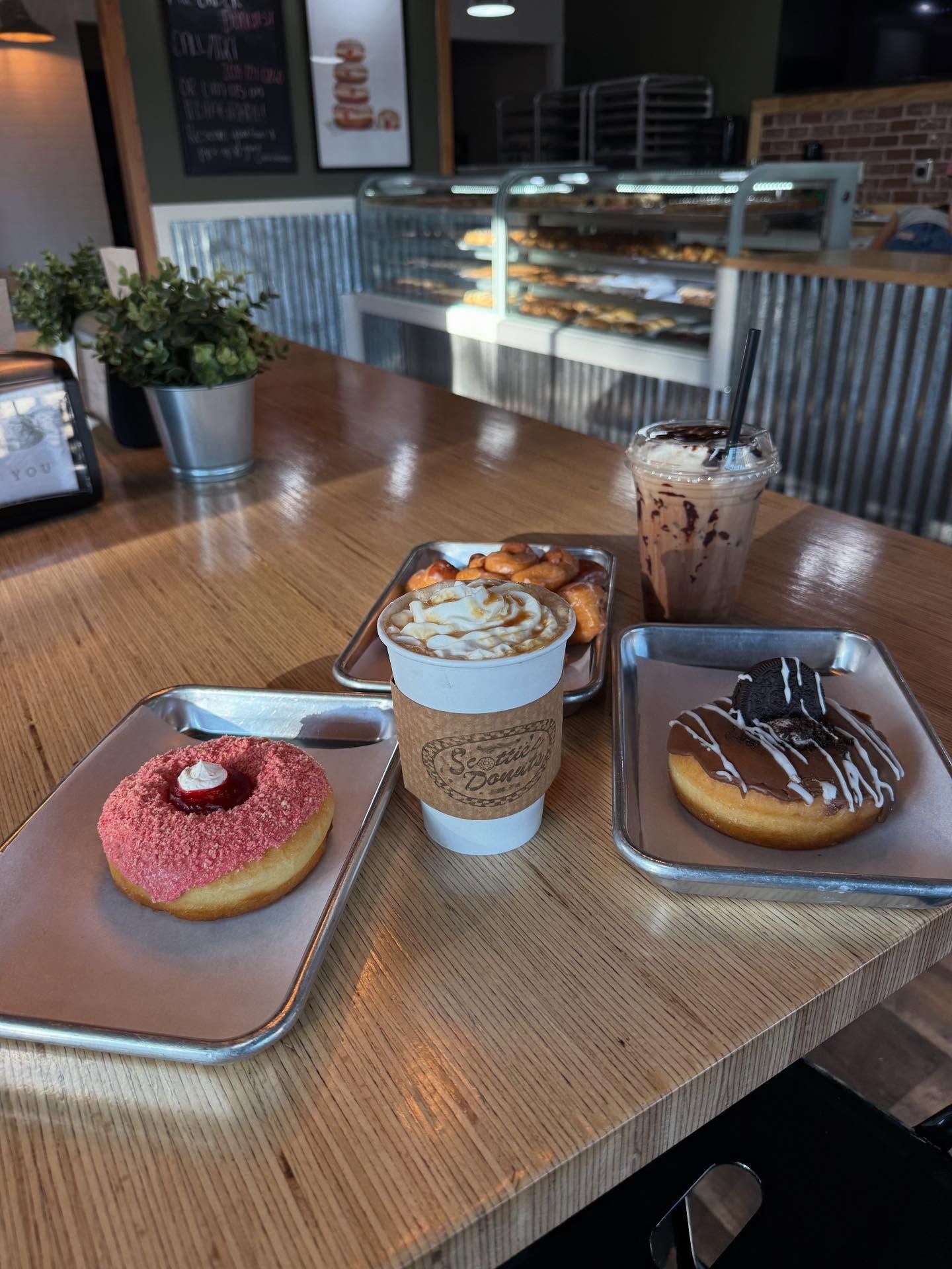 Donuts and drinks on a wooden table in a bakery. A pink donut, coffee, and a chocolate donut are visible.