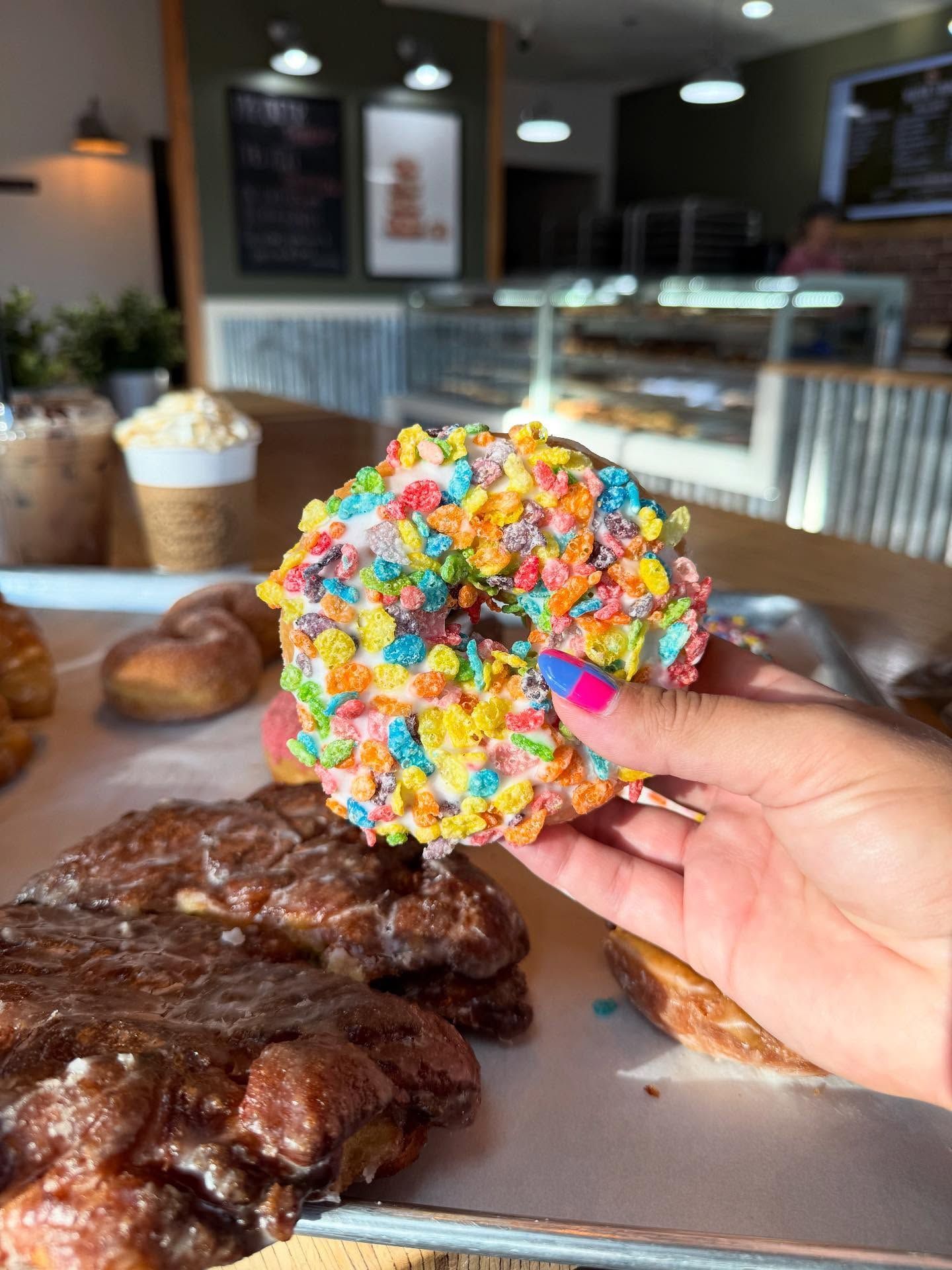 A hand holding a colorful cereal-covered donut in a bakery.