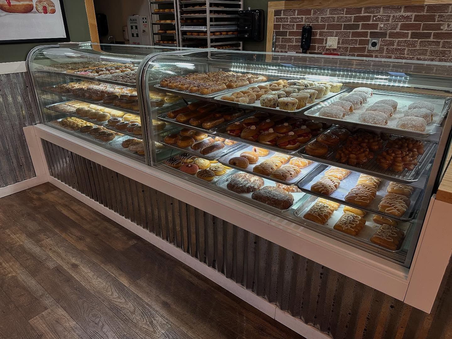 Donut display case at a bakery with rows of various donuts lit by interior lights. Wooden floor and wood-paneled walls surround.