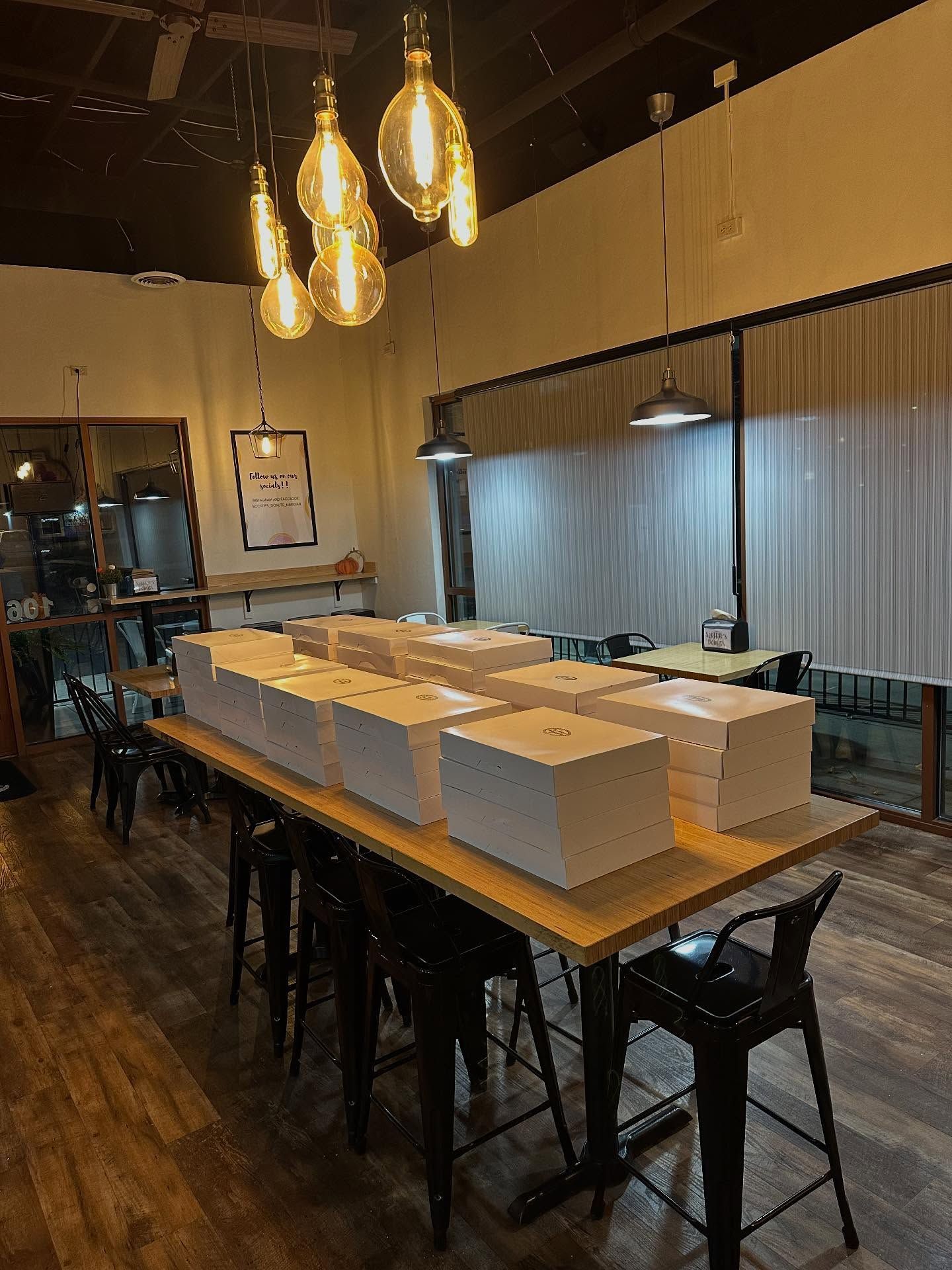 Large table with boxes and chairs in a cafe, illuminated by hanging lights, with a window and a side door visible.