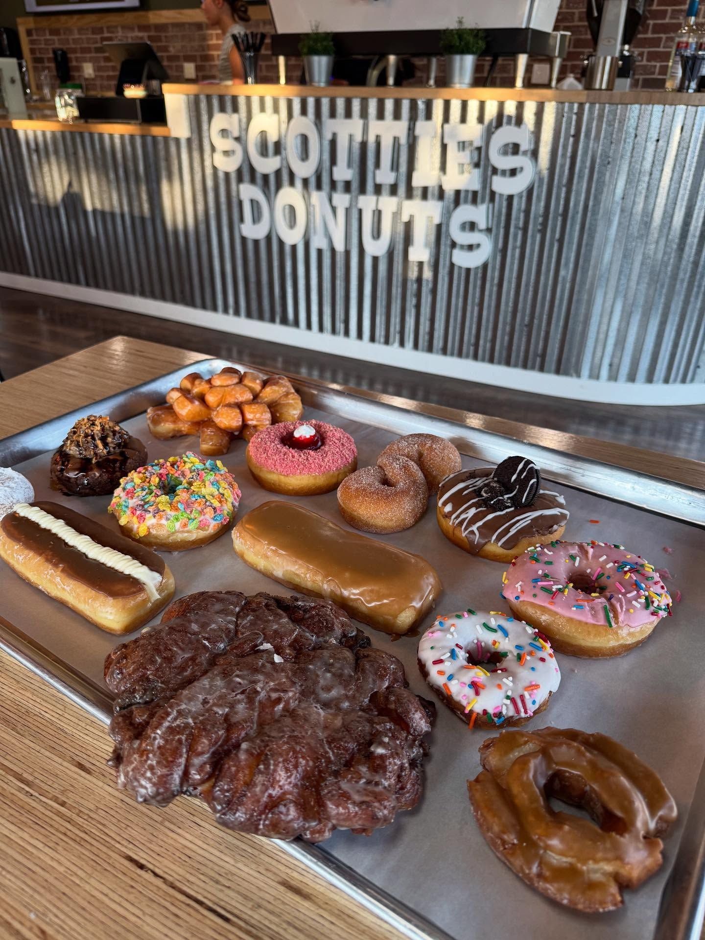 A tray of various glazed donuts from Scottie's Donuts, including chocolate, sprinkles, and a large apple fritter.