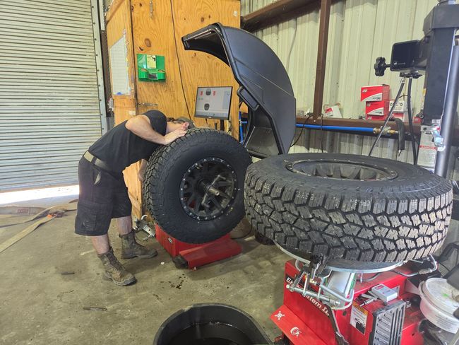 Mechanic in workshop changing a large truck tire beside mounted wheels and red tire-changing equipment | Phase 2 Automotive
