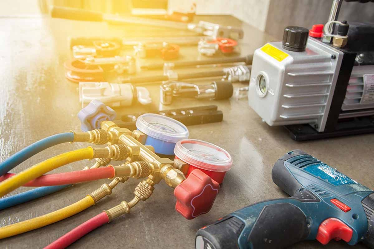HVAC tools: gauges, hoses, vacuum pump, and power drill on a light-colored surface, likely for air conditioning repair.
