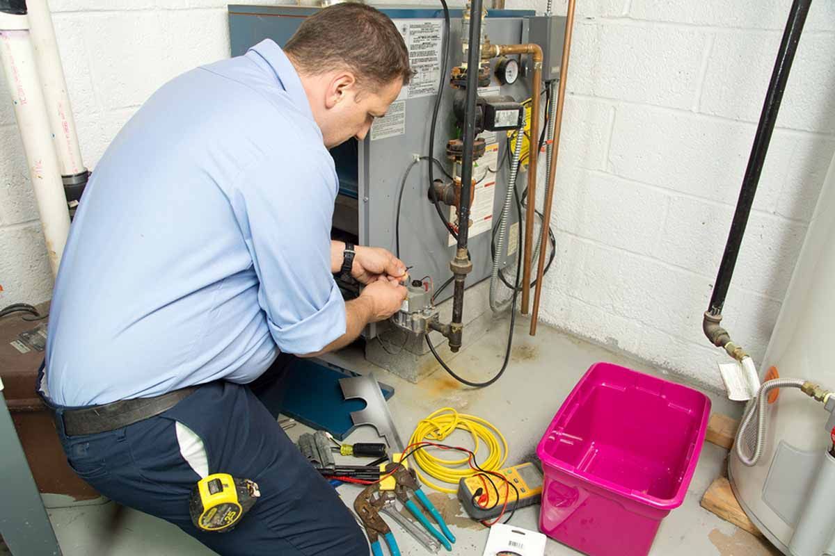 HVAC technician in blue shirt repairing furnace in a basement, tools and pink bin on the floor.