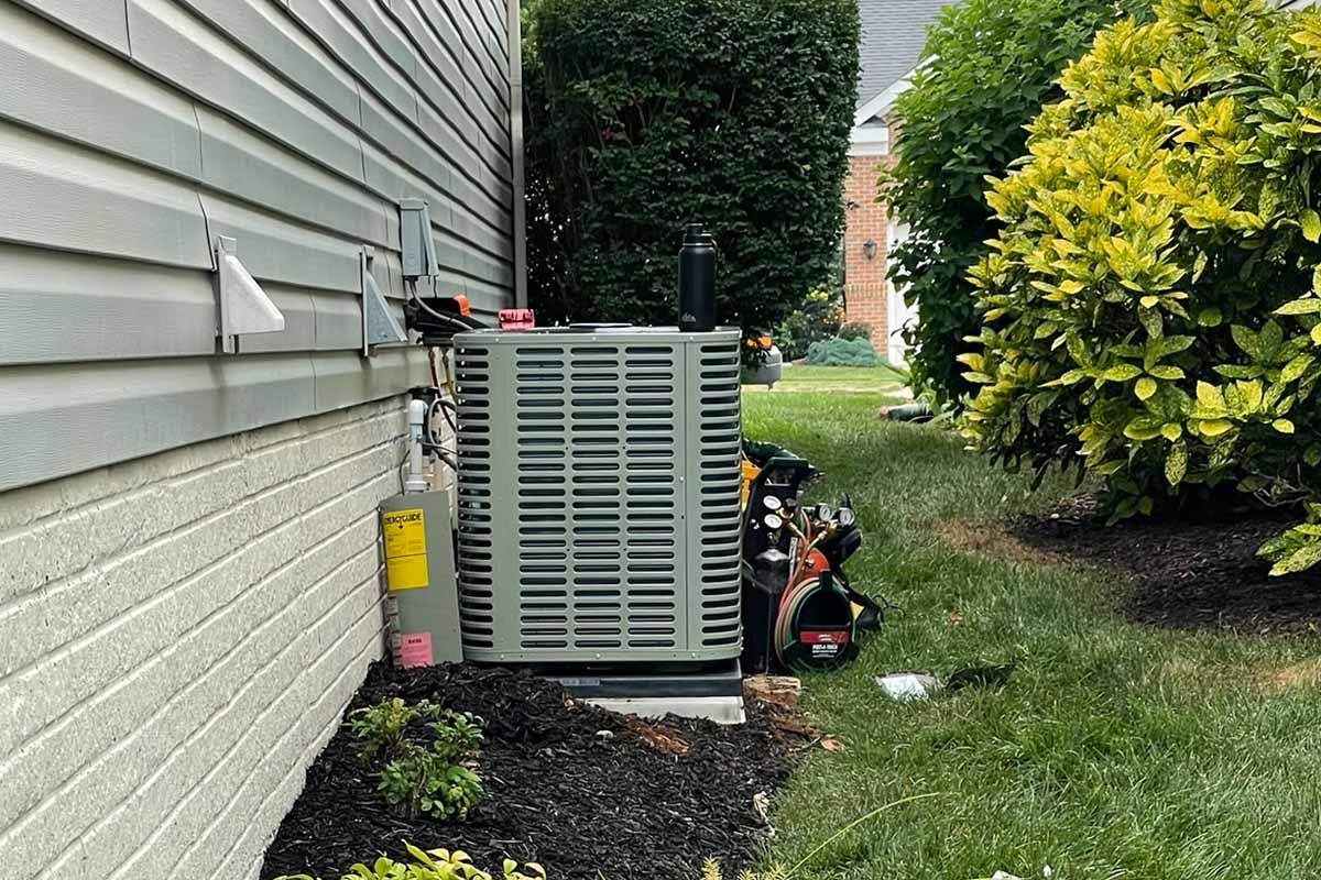 An outdoor air conditioning unit against a light gray house with greenery surrounding it.