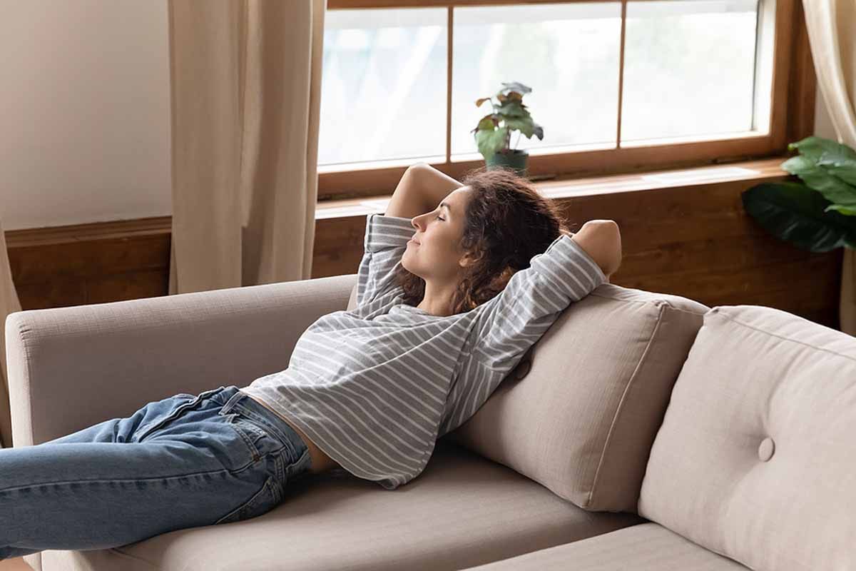 Woman relaxing on a couch, arms behind head, eyes closed, near a window with a plant.