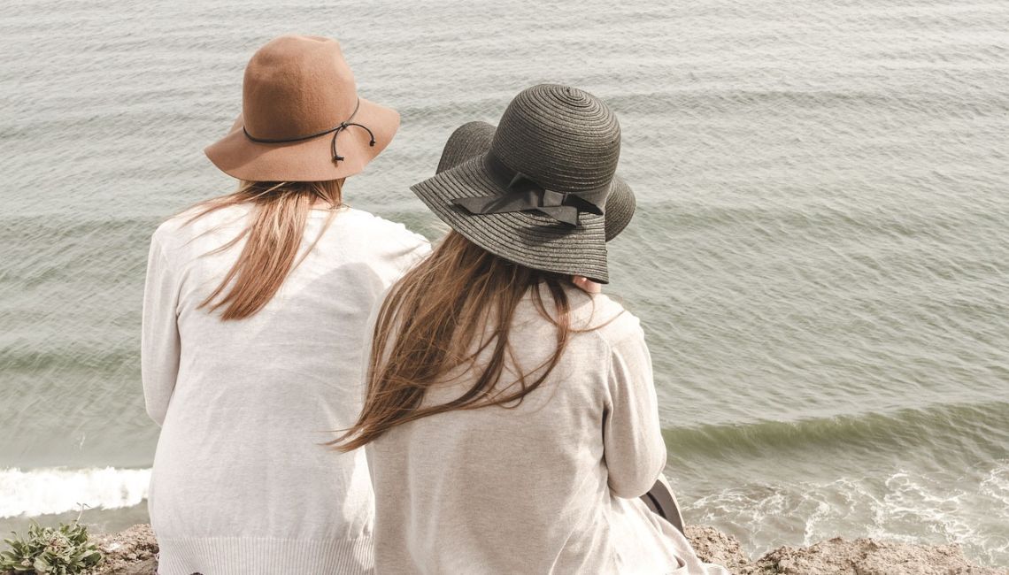 Dos mujeres con sombreros sentadas de espaldas, contemplando el mar desde un acantilado