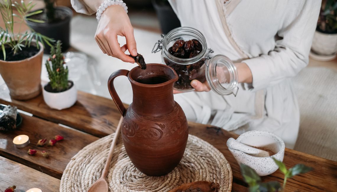 Mujer preparando una ceremonia de cacao: coloca un dátil en una jarra de barro tallada, rodeada de velas, plantas y utensilios artesanales sobre una mesa de madera.