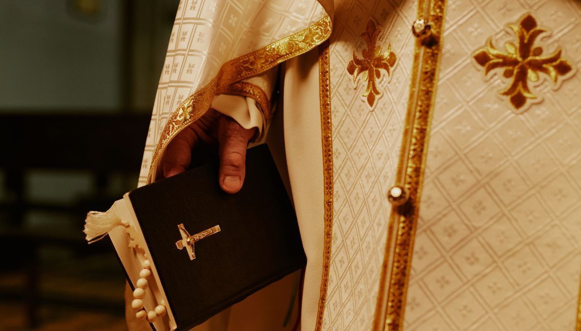 Un sacerdote sostiene una Biblia negra con un crucifijo dorado, vestido con una túnica litúrgica blanca y dorada decorada con cruces.