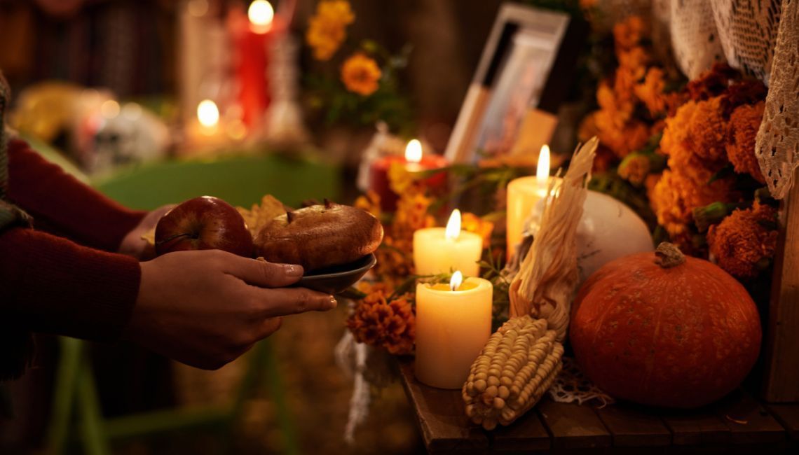 Manos colocando manzana y pan en una ofrenda con velas, cempasúchil, calabaza y maí