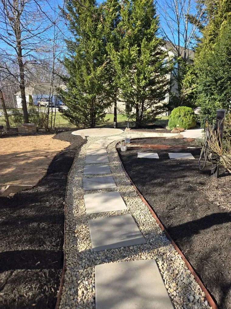 Curved stone walkway through a landscaped yard with trees and a raised garden bed under a blue sky