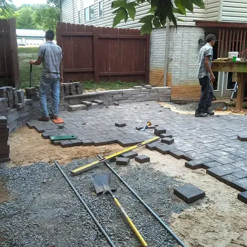 Two workers laying gray pavers in a backyard patio, with tools and gravel in the foreground.