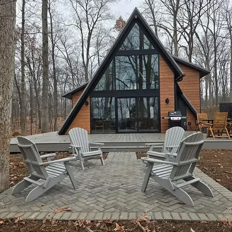 A-frame cabin with large windows and wooden deck, surrounded by leafless trees and outdoor lounge chairs.